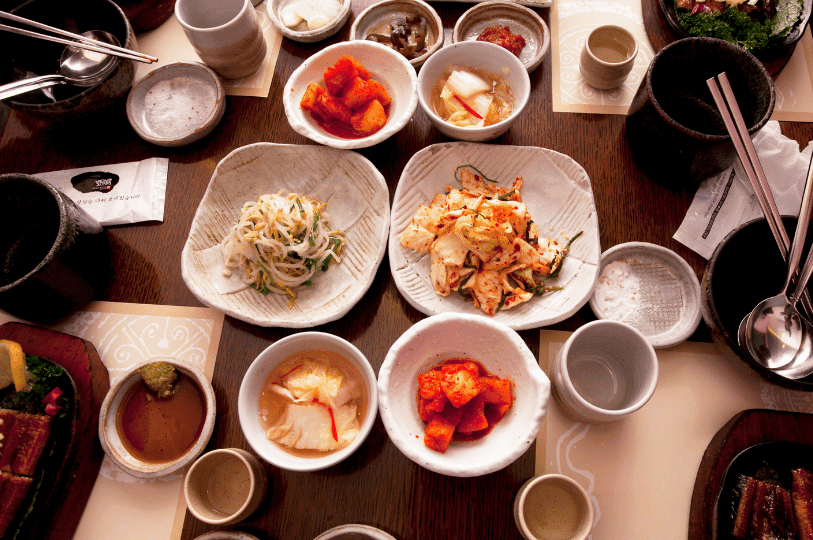 A rustic, overhead view of a wooden table richly crowded with an array of banchan in artisanal ceramic bowls. The abundant spread of Napa cabbage kimchi, bean sprouts, and radish kimchi reflects the communal and generous spirit of Korean dining. A rustic, overhead view of a wooden table richly crowded with an array of banchan in artisanal ceramic bowls. The abundant spread of Napa cabbage kimchi, bean sprouts, and radish kimchi reflects the communal and generous spirit of Korean dining.