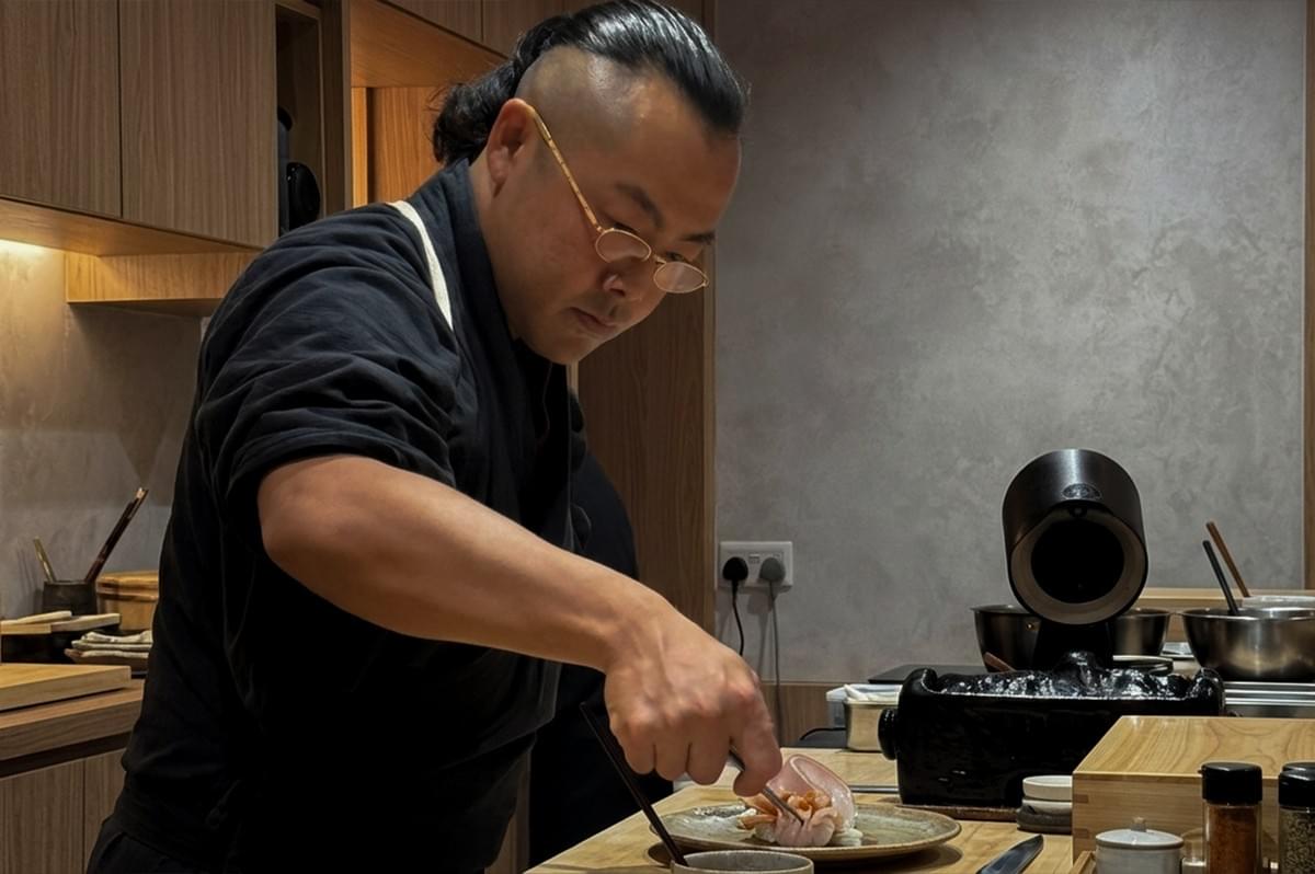 A medium side-profile shot captures Chef Masa, a man with a shaved head except for a dark ponytail bun, wearing thin, gold-rimmed glasses and a dark grey chef's uniform. He is leaning forward over a wooden countertop in a modern, minimalist kitchen, focusing intently on plating a dish. With his right hand, he uses fine tweezers to delicately arrange thin slices of pale pink food, likely seafood or meat, on a round ceramic plate, while holding a pair of chopsticks in his left hand. The background features light-toned wooden cabinetry, textured grey walls, an electrical outlet, and various kitchen tools, including stainless steel bowls and spice shakers, all contributing to a clean and professional culinary setting. A medium side-profile shot captures Chef Masa, a man with a shaved head except for a dark ponytail bun, wearing thin, gold-rimmed glasses and a dark grey chef's uniform. He is leaning forward over a wooden countertop in a modern, minimalist kitchen, focusing intently on plating a dish. With his right hand, he uses fine tweezers to delicately arrange thin slices of pale pink food, likely seafood or meat, on a round ceramic plate, while holding a pair of chopsticks in his left hand. The background features light-toned wooden cabinetry, textured grey walls, an electrical outlet, and various kitchen tools, including stainless steel bowls and spice shakers, all contributing to a clean and professional culinary setting.
