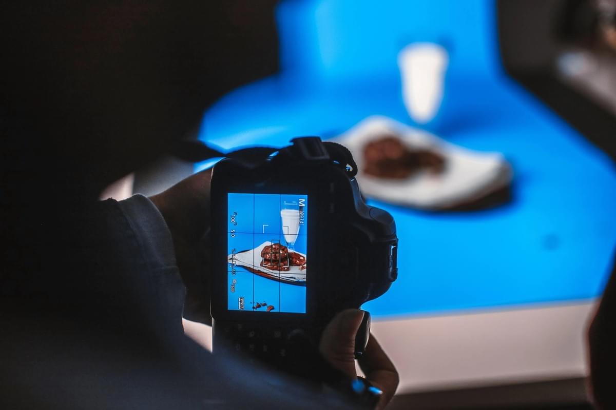 A photographer holds a DSLR camera displaying a shot of chocolate cookies and a glass of milk on its LCD screen. In the background, the actual food setup sits arranged on a bright blue backdrop inside a studio setting. A photographer holds a DSLR camera displaying a shot of chocolate cookies and a glass of milk on its LCD screen. In the background, the actual food setup sits arranged on a bright blue backdrop inside a studio setting.