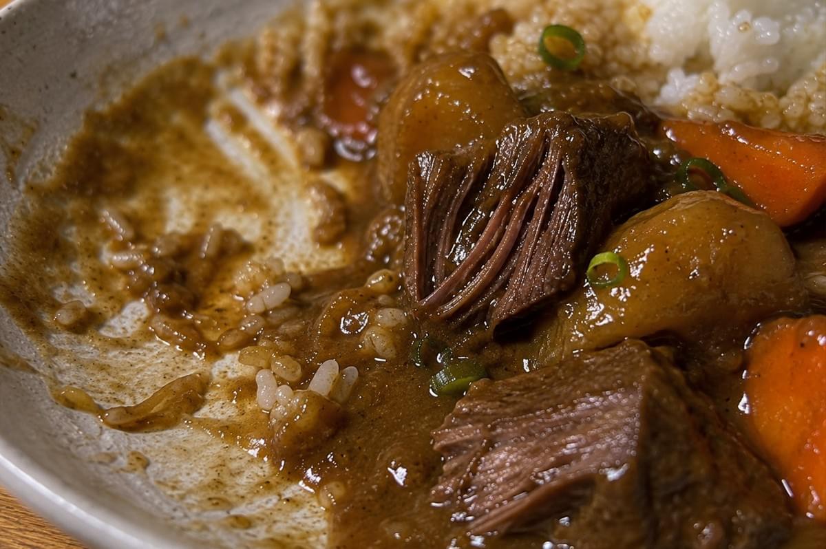 An extreme close-up macro shot of a partially eaten plate of curry, showcasing the incredibly tender texture of the slow-braised beef. One large piece of beef has been pulled apart, clearly revealing the succulent, juicy, and perfectly separated meat fibers coated in the glossy, thick brown curry sauce. Soft-cooked carrots and potatoes are nestled nearby, with a few grains of white rice clinging to the rich sauce on the side of the ceramic dish. An extreme close-up macro shot of a partially eaten plate of curry, showcasing the incredibly tender texture of the slow-braised beef. One large piece of beef has been pulled apart, clearly revealing the succulent, juicy, and perfectly separated meat fibers coated in the glossy, thick brown curry sauce. Soft-cooked carrots and potatoes are nestled nearby, with a few grains of white rice clinging to the rich sauce on the side of the ceramic dish.
