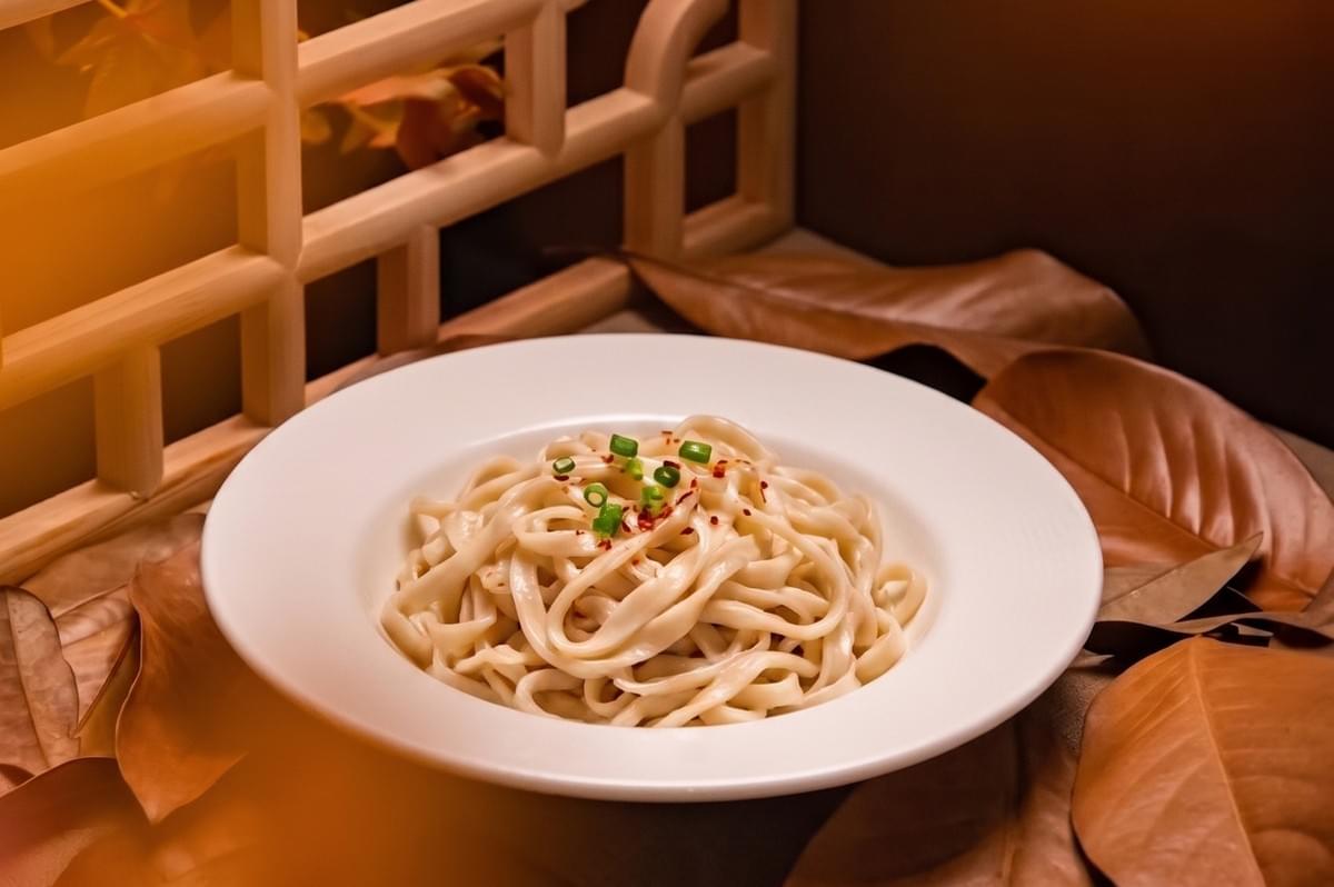 White bowl of sesame noodles garnished with green onions and chili flakes in an autumn-themed setting. White bowl of sesame noodles garnished with green onions and chili flakes in an autumn-themed setting.