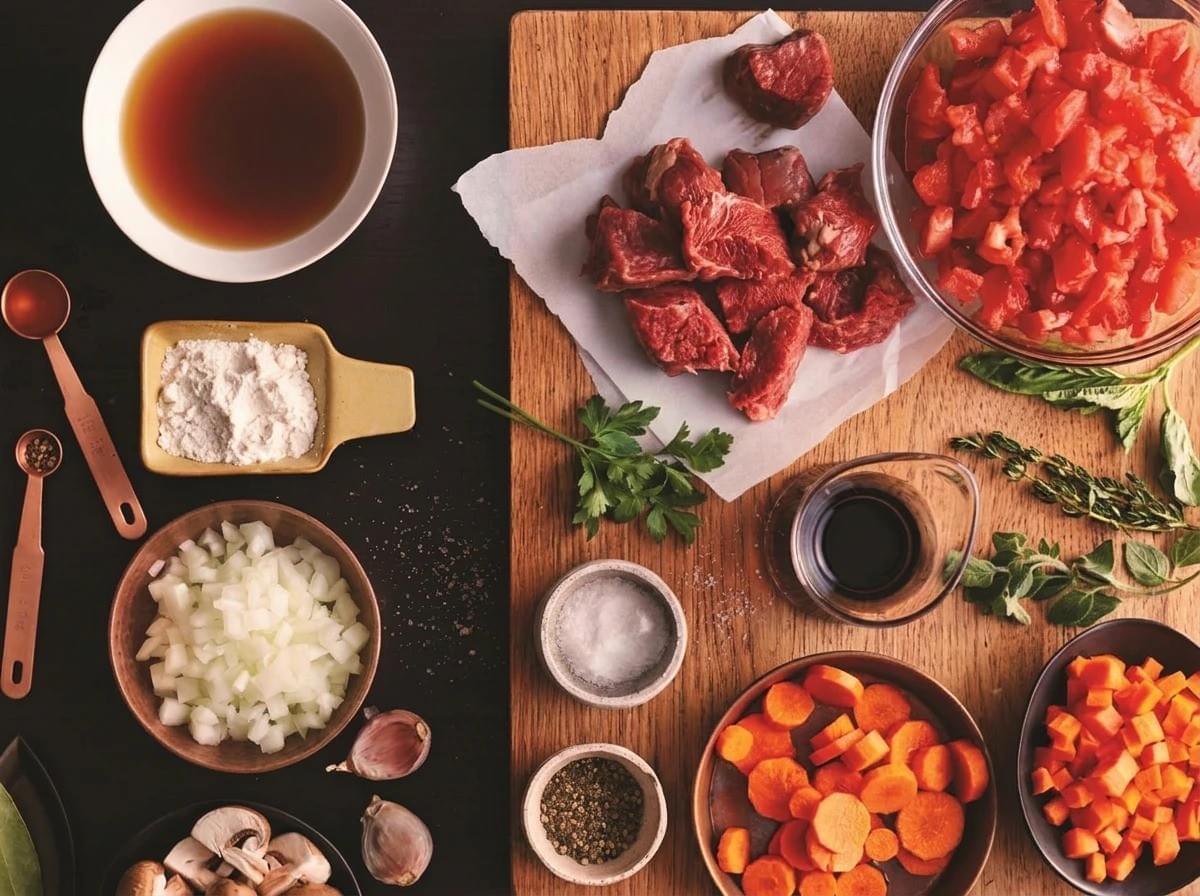 An overhead, split-background shot showing prepped ingredients for a stew, featuring raw cubed beef on a wooden cutting board alongside diced tomatoes, carrots, herbs, broth, chopped onions, and spices. An overhead, split-background shot showing prepped ingredients for a stew, featuring raw cubed beef on a wooden cutting board alongside diced tomatoes, carrots, herbs, broth, chopped onions, and spices.