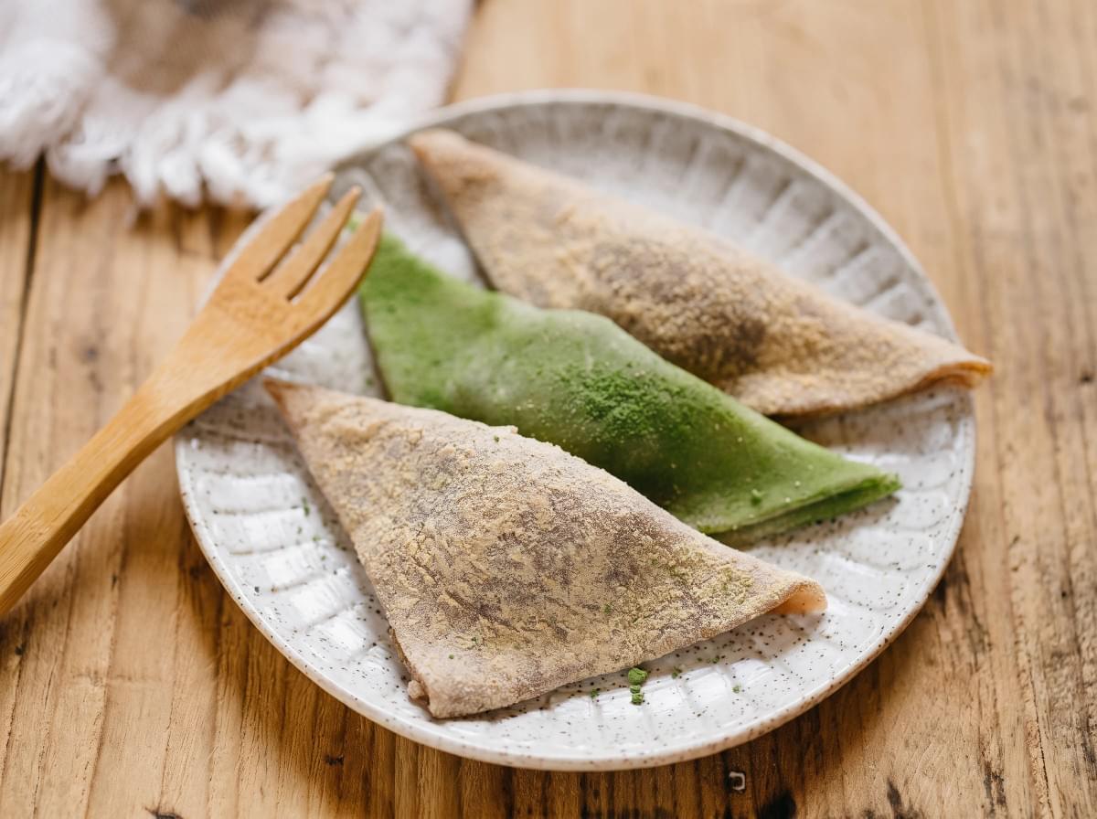 Three dusted triangular yatsuhashi mochi pieces on a speckled ceramic plate with a wooden fork. Three dusted triangular yatsuhashi mochi pieces on a speckled ceramic plate with a wooden fork.