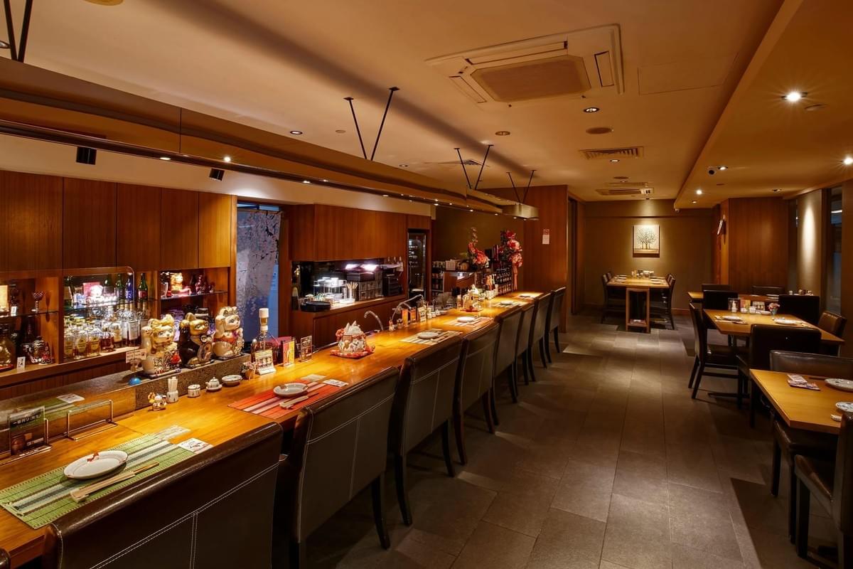 A long, warm-toned wooden bar counter with leather chairs stretches down the left side of a dimly lit Japanese restaurant, decorated with maneki-neko figures and liquor bottles. To the right, several small wooden dining tables and chairs are neatly arranged across the tiled floor under soft recessed ceiling lights. A long, warm-toned wooden bar counter with leather chairs stretches down the left side of a dimly lit Japanese restaurant, decorated with maneki-neko figures and liquor bottles. To the right, several small wooden dining tables and chairs are neatly arranged across the tiled floor under soft recessed ceiling lights.