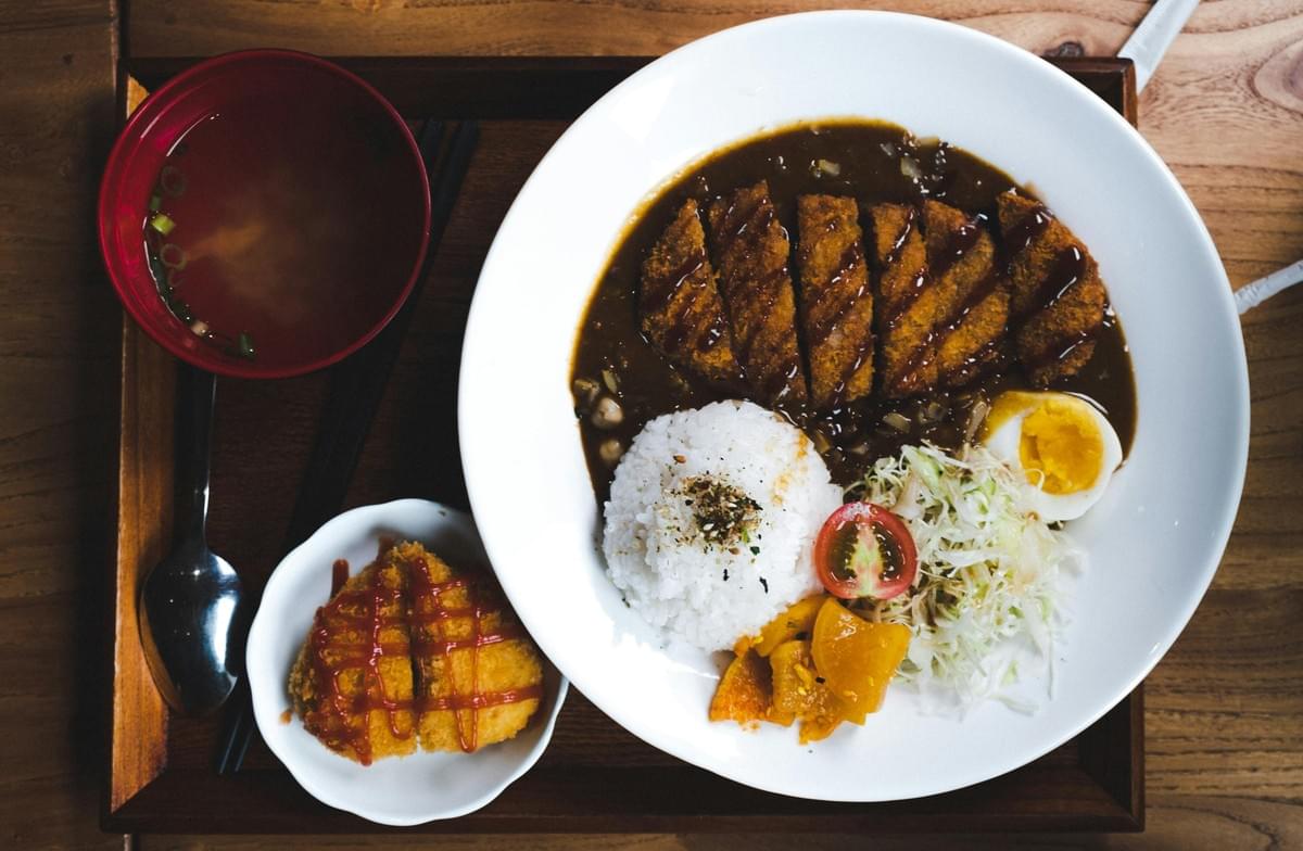 A high-angle shot shows a Japanese chicken katsu curry meal beautifully arranged on a wooden tray. The main white plate features sliced breaded cutlets over rich curry sauce, a scoop of white rice, shredded cabbage salad with a tomato slice, half a boiled egg, and pickled vegetables, accompanied by a small side bowl of miso soup and an extra fried cutlet drizzled with ketchup. A high-angle shot shows a Japanese chicken katsu curry meal beautifully arranged on a wooden tray. The main white plate features sliced breaded cutlets over rich curry sauce, a scoop of white rice, shredded cabbage salad with a tomato slice, half a boiled egg, and pickled vegetables, accompanied by a small side bowl of miso soup and an extra fried cutlet drizzled with ketchup.
