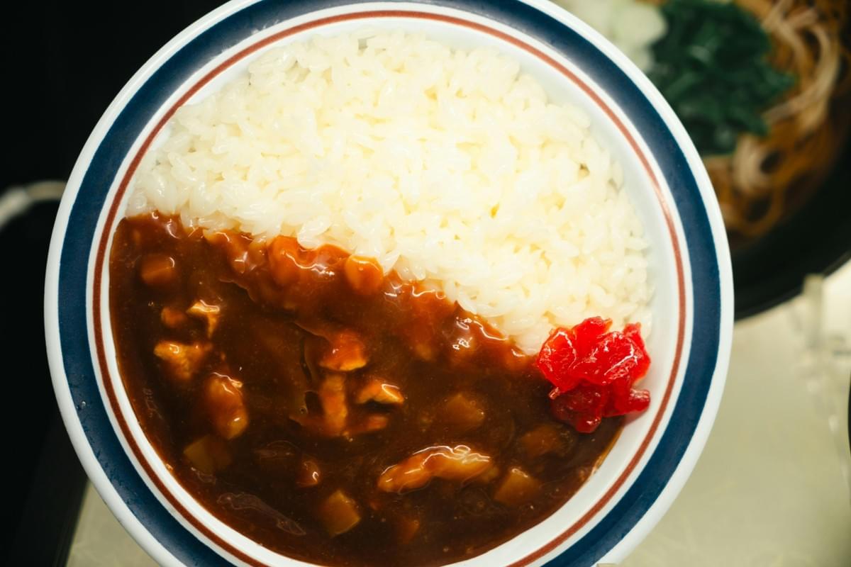 A top-down view of a round white bowl containing Japanese curry rice, with fluffy white rice filling the top half and a thick brown curry sauce with visible chunks of meat and vegetables covering the bottom half. A small serving of bright red pickled radish (fukujinzuke) sits on the side where the rice and curry meet. A top-down view of a round white bowl containing Japanese curry rice, with fluffy white rice filling the top half and a thick brown curry sauce with visible chunks of meat and vegetables covering the bottom half. A small serving of bright red pickled radish (fukujinzuke) sits on the side where the rice and curry meet.