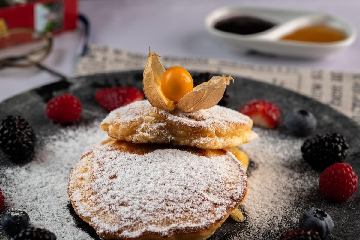 A stack of fluffy pancakes heavily dusted with powdered sugar sits on a dark plate, topped with a single cape gooseberry in its papery husk. The dish is surrounded by scattered fresh berries, including raspberries, blueberries, and blackberries, with syrups visible in the blurred background. A stack of fluffy pancakes heavily dusted with powdered sugar sits on a dark plate, topped with a single cape gooseberry in its papery husk. The dish is surrounded by scattered fresh berries, including raspberries, blueberries, and blackberries, with syrups visible in the blurred background.