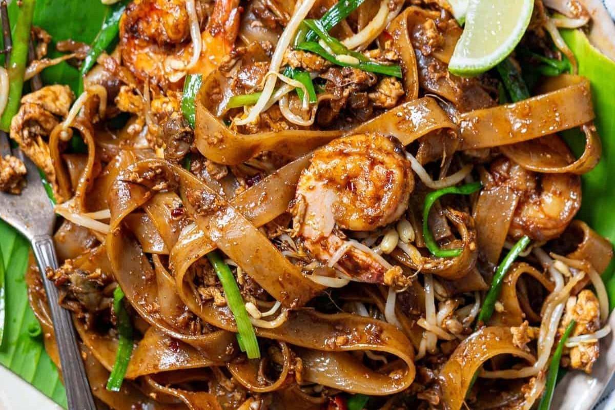 This image features a close-up of Char Kway Teow, a Southeast Asian stir-fried noodle dish served on a green banana leaf. It showcases flat rice noodles tossed with shrimp, bean sprouts, green chives, and egg in a rich, dark sauce. This image features a close-up of Char Kway Teow, a Southeast Asian stir-fried noodle dish served on a green banana leaf. It showcases flat rice noodles tossed with shrimp, bean sprouts, green chives, and egg in a rich, dark sauce.