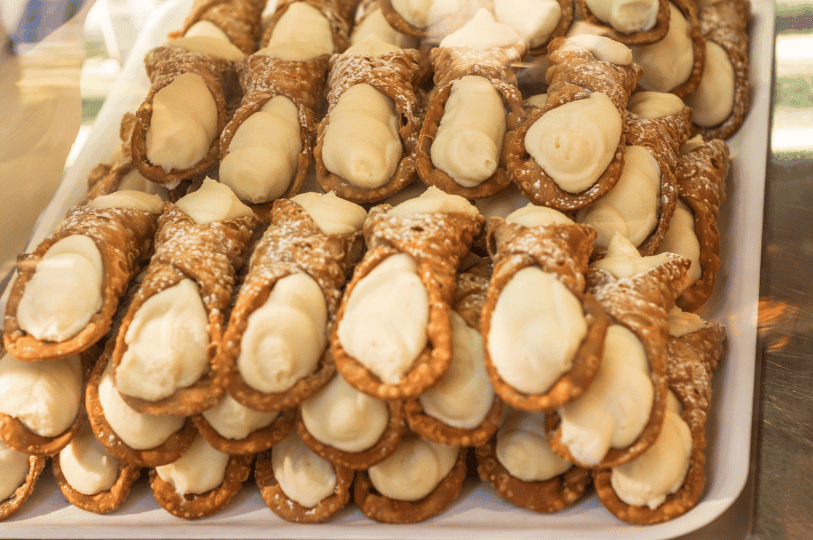 A large tray of freshly filled cannoli stacked in rows, showing off the bubbly, fried texture of the pastry shells and the generous amount of creamy white filling. A large tray of freshly filled cannoli stacked in rows, showing off the bubbly, fried texture of the pastry shells and the generous amount of creamy white filling.