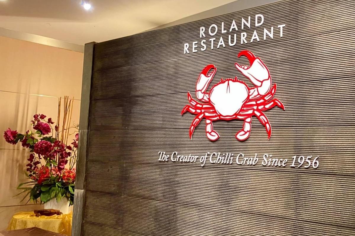 A dark slatted wall features white lettering for "ROLAND RESTAURANT" positioned above a prominent red and white crab logo. Beneath the crab, white script text reads "The Creator of Chilli Crab Since 1956," with a floral arrangement visible in the background to the left. A dark slatted wall features white lettering for "ROLAND RESTAURANT" positioned above a prominent red and white crab logo. Beneath the crab, white script text reads "The Creator of Chilli Crab Since 1956," with a floral arrangement visible in the background to the left.