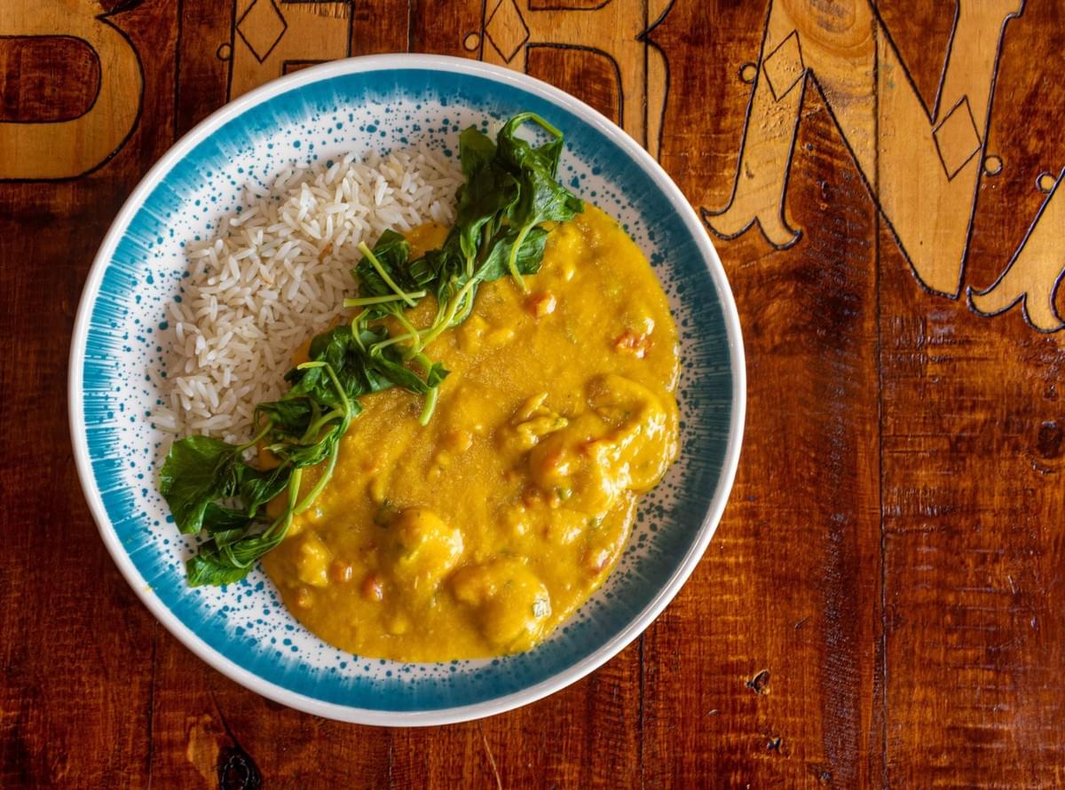 A top-down view of a round, blue-rimmed plate featuring rich yellow curry alongside white basmati rice, topped with fresh green herbs. The plate rests on a warm, rustic wooden tabletop with carved lettering. A top-down view of a round, blue-rimmed plate featuring rich yellow curry alongside white basmati rice, topped with fresh green herbs. The plate rests on a warm, rustic wooden tabletop with carved lettering.