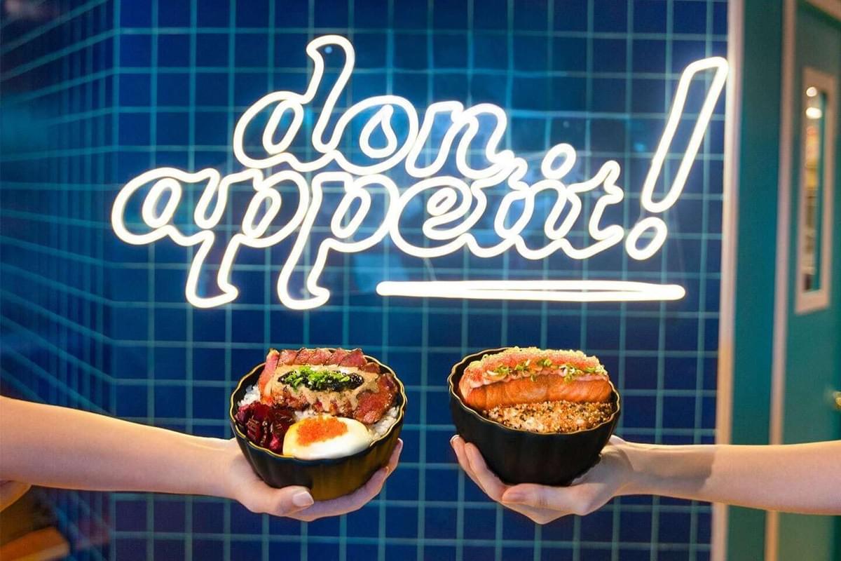 Two hands hold up dark ceramic bowls of rice topped with sliced beef and seared salmon. In the background, a glowing white neon sign reads "don appetit!" against a blue tiled wall. Two hands hold up dark ceramic bowls of rice topped with sliced beef and seared salmon. In the background, a glowing white neon sign reads "don appetit!" against a blue tiled wall.