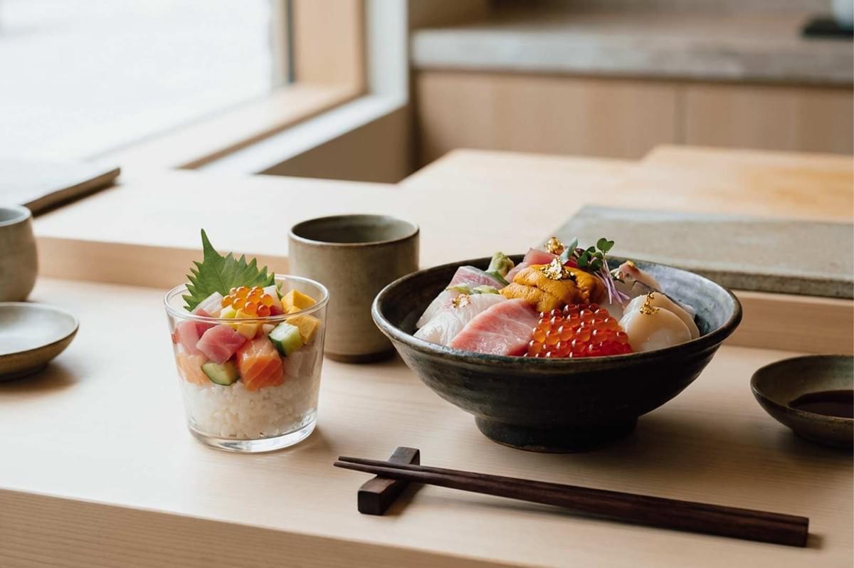 An elegant Japanese dining setup featuring a premium dark ceramic chirashi bowl generously filled with fresh sashimi, sea urchin, ikura, scallops, and gold leaf garnishes, positioned beside a clear glass containing layered bara chirashi cubes over rice, set with wooden chopsticks and ceramic teaware on a light minimalist wooden counter. An elegant Japanese dining setup featuring a premium dark ceramic chirashi bowl generously filled with fresh sashimi, sea urchin, ikura, scallops, and gold leaf garnishes, positioned beside a clear glass containing layered bara chirashi cubes over rice, set with wooden chopsticks and ceramic teaware on a light minimalist wooden counter.