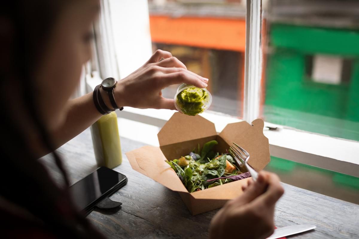 A woman pours green dressing from a small container onto a fresh salad in a takeout box. She sits near a window with a fork in hand, next to a smartphone resting on the wooden counter. A woman pours green dressing from a small container onto a fresh salad in a takeout box. She sits near a window with a fork in hand, next to a smartphone resting on the wooden counter.