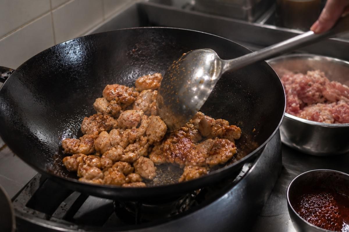 A black wok sits over a stove flame as a metal ladle tosses bite-sized pieces of seasoned chicken, creating a light sear as they cook in oil. Next to the wok, a metal prep bowl contains additional raw seasoned meat, and a small container of red sambal rests nearby on the stainless steel surface. A black wok sits over a stove flame as a metal ladle tosses bite-sized pieces of seasoned chicken, creating a light sear as they cook in oil. Next to the wok, a metal prep bowl contains additional raw seasoned meat, and a small container of red sambal rests nearby on the stainless steel surface.