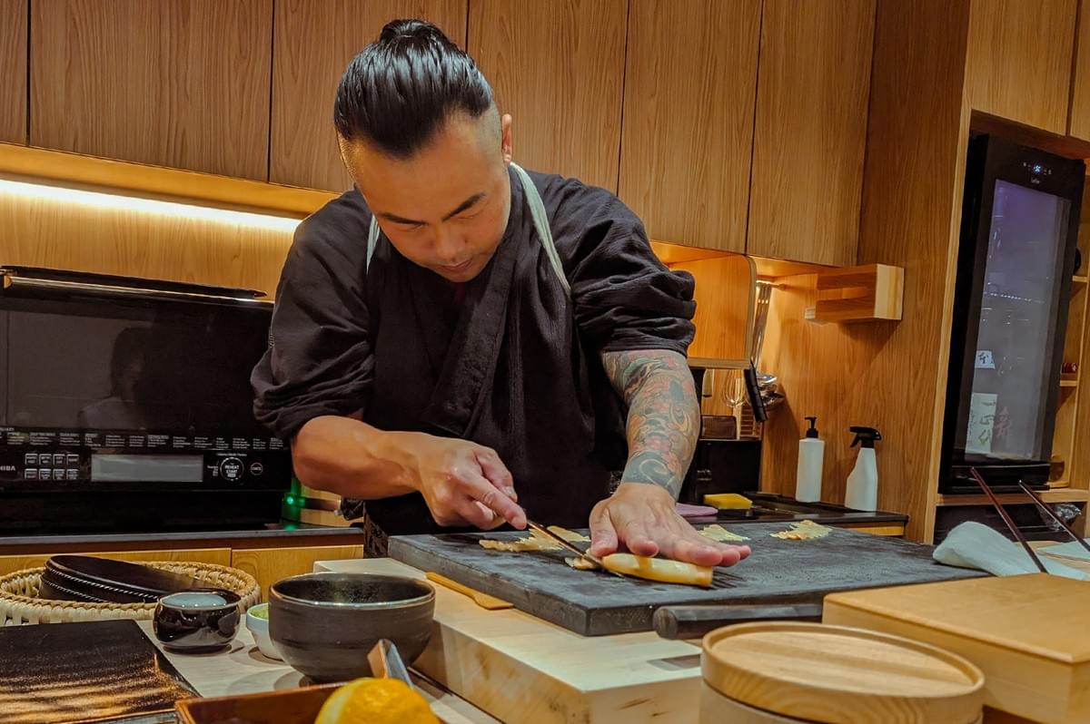 Chef preparing ingredients on a wooden cutting board inside a warm, wood-accented omakase kitchen. Chef preparing ingredients on a wooden cutting board inside a warm, wood-accented omakase kitchen.