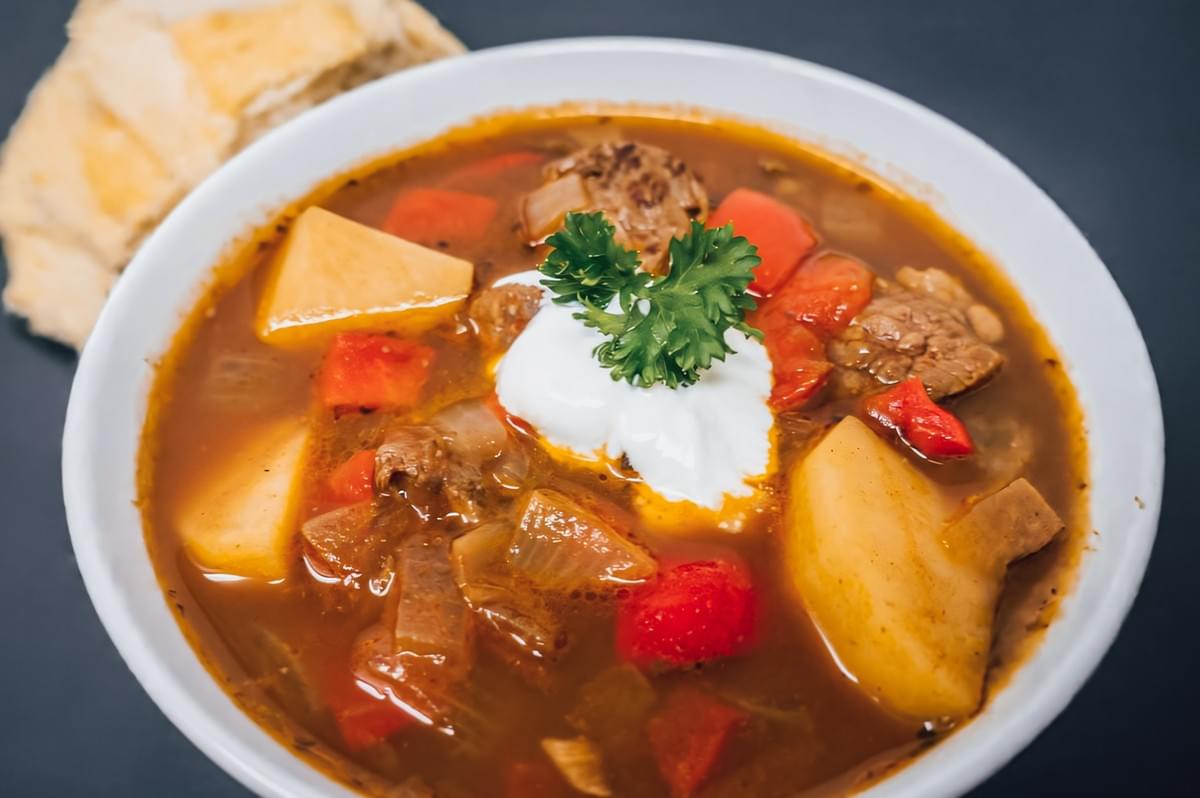 Overhead top-down shot of German-style beef goulash soup with potatoes, vegetables, and sour cream garnish, served with bread, showcasing warm hearty comfort food in a tropical climate adaptation context Overhead top-down shot of German-style beef goulash soup with potatoes, vegetables, and sour cream garnish, served with bread, showcasing warm hearty comfort food in a tropical climate adaptation context