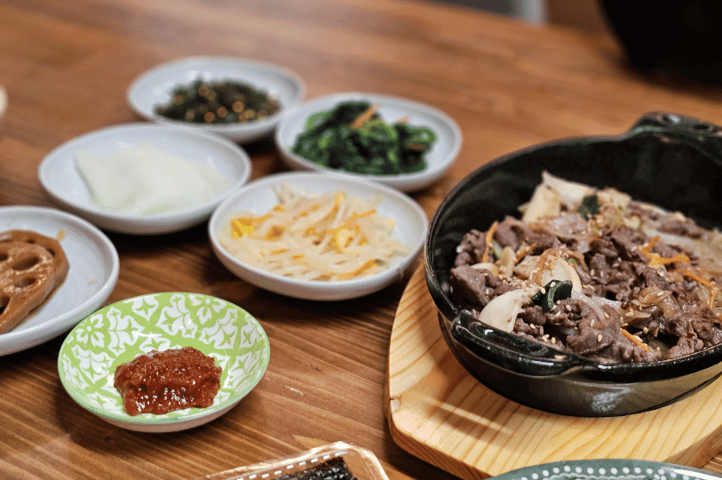 A warm, angled shot focusing on the harmonious pairing of a hearty beef bulgogi skillet with essential vegetable banchan. Small dishes of seasoned spinach, bean sprouts, pickled radish, and braised lotus root frame the main dish. A warm, angled shot focusing on the harmonious pairing of a hearty beef bulgogi skillet with essential vegetable banchan. Small dishes of seasoned spinach, bean sprouts, pickled radish, and braised lotus root frame the main dish.