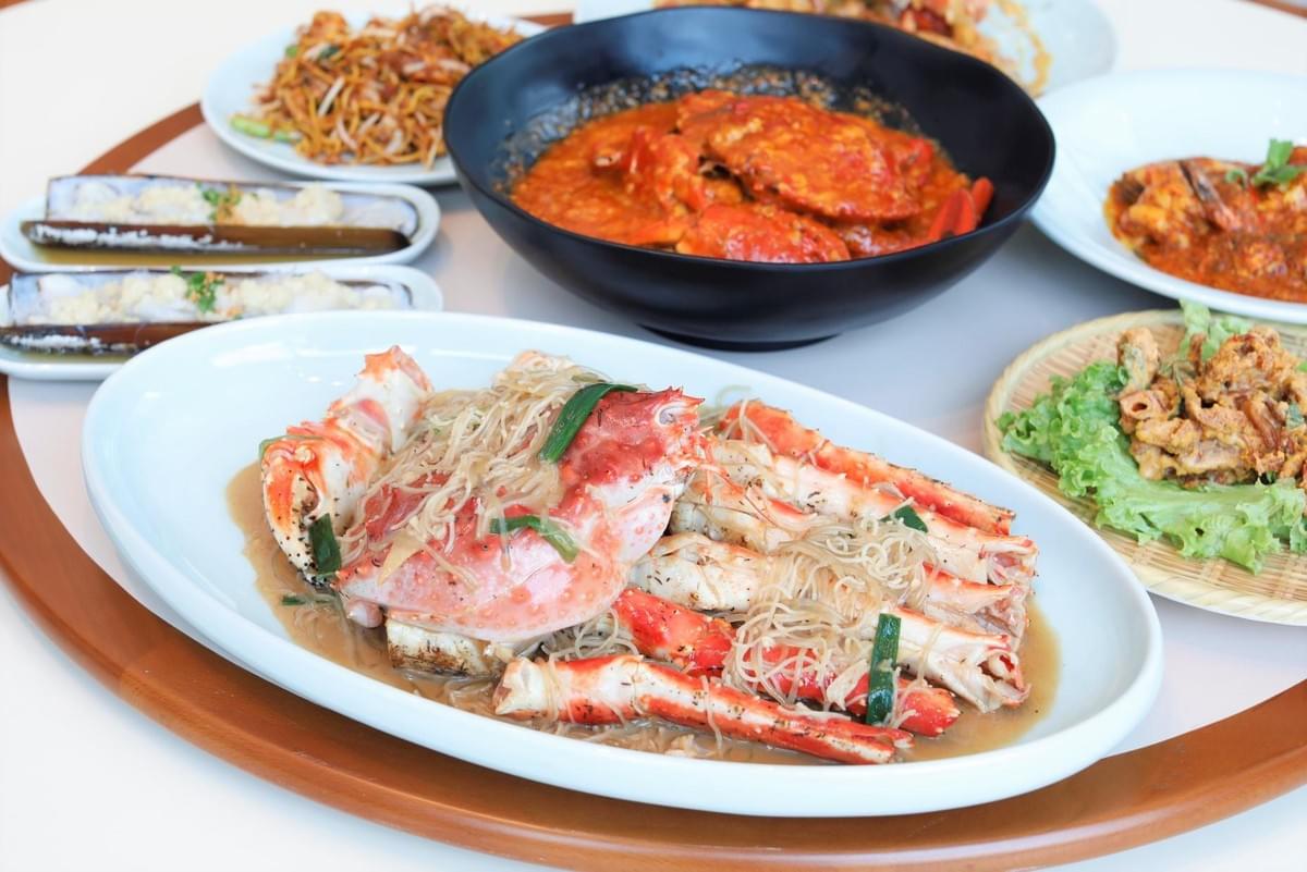 A round dining table is filled with a rich spread of seafood dishes, featuring a large oval platter of steamed king crab legs with glass noodles upfront. Surrounding it are bowls and plates of chilli crab, stir-fried noodles, steamed bamboo clams, and crispy fried appetizers. A round dining table is filled with a rich spread of seafood dishes, featuring a large oval platter of steamed king crab legs with glass noodles upfront. Surrounding it are bowls and plates of chilli crab, stir-fried noodles, steamed bamboo clams, and crispy fried appetizers.