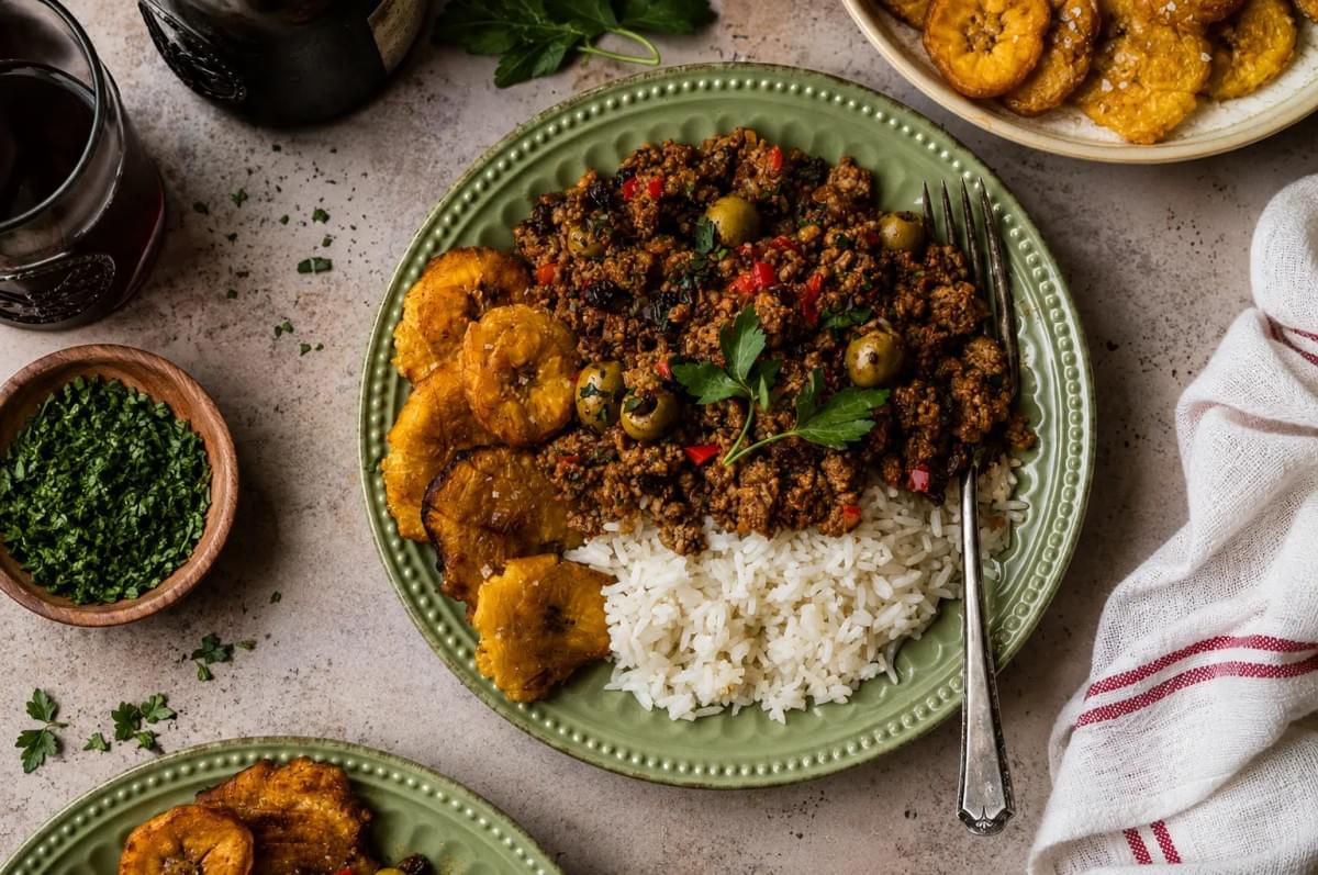 Overhead view of a green plate featuring picadillo mixed with green olives, served alongside white rice and several golden-brown fried plantains. Overhead view of a green plate featuring picadillo mixed with green olives, served alongside white rice and several golden-brown fried plantains.