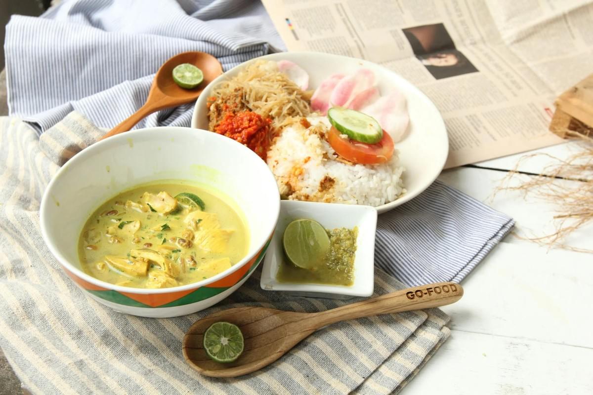 In the image, a vibrant spread of Indonesian cuisine is arranged on a striped cloth next to a newspaper. The meal features a bowl of creamy yellow chicken curry (Opor Ayam) accompanied by a plate of white rice, noodles, sambal, and colorful crackers. In the image, a vibrant spread of Indonesian cuisine is arranged on a striped cloth next to a newspaper. The meal features a bowl of creamy yellow chicken curry (Opor Ayam) accompanied by a plate of white rice, noodles, sambal, and colorful crackers.