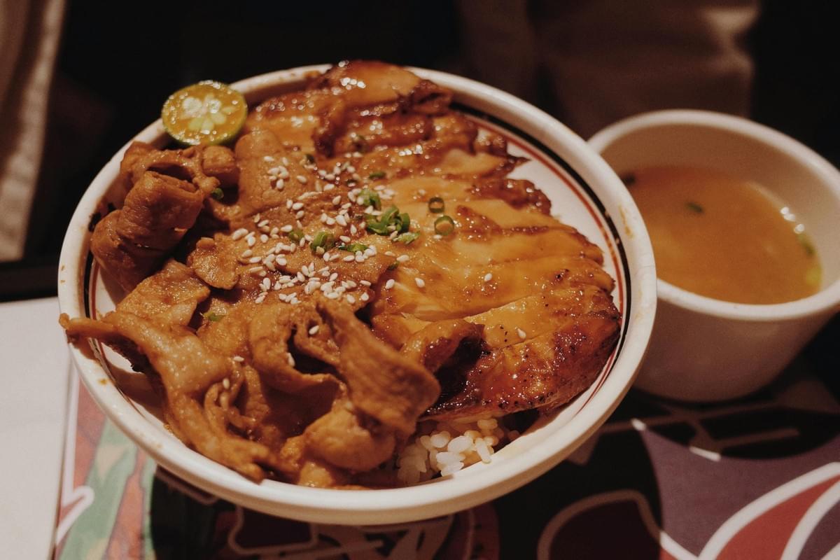 A close-up shot of a rice bowl topped with glazed meats, sesame seeds, green onions, and a small citrus slice on the rim. Next to the main dish, a small white bowl filled with light broth sits on the table. A close-up shot of a rice bowl topped with glazed meats, sesame seeds, green onions, and a small citrus slice on the rim. Next to the main dish, a small white bowl filled with light broth sits on the table.