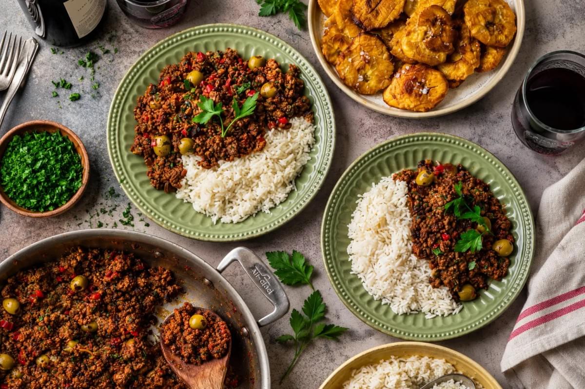 Flat lay of a dinner table setting with two green plates of picadillo and rice, a large frying pan, a side dish of fried plantains, and glasses of red wine. Flat lay of a dinner table setting with two green plates of picadillo and rice, a large frying pan, a side dish of fried plantains, and glasses of red wine.