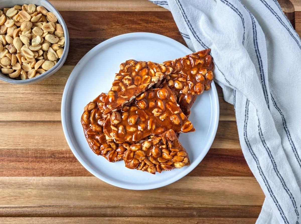 An overhead view of a white plate stacked with glossy, dark-amber peanut brittle on a wooden board, next to a small bowl of roasted peanuts and a striped kitchen towel. An overhead view of a white plate stacked with glossy, dark-amber peanut brittle on a wooden board, next to a small bowl of roasted peanuts and a striped kitchen towel.
