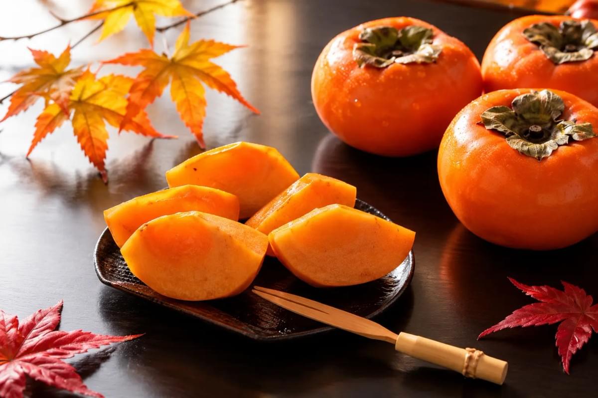 Fresh persimmon slices served on a small dark plate alongside whole persimmons and autumn maple leaves. Fresh persimmon slices served on a small dark plate alongside whole persimmons and autumn maple leaves.