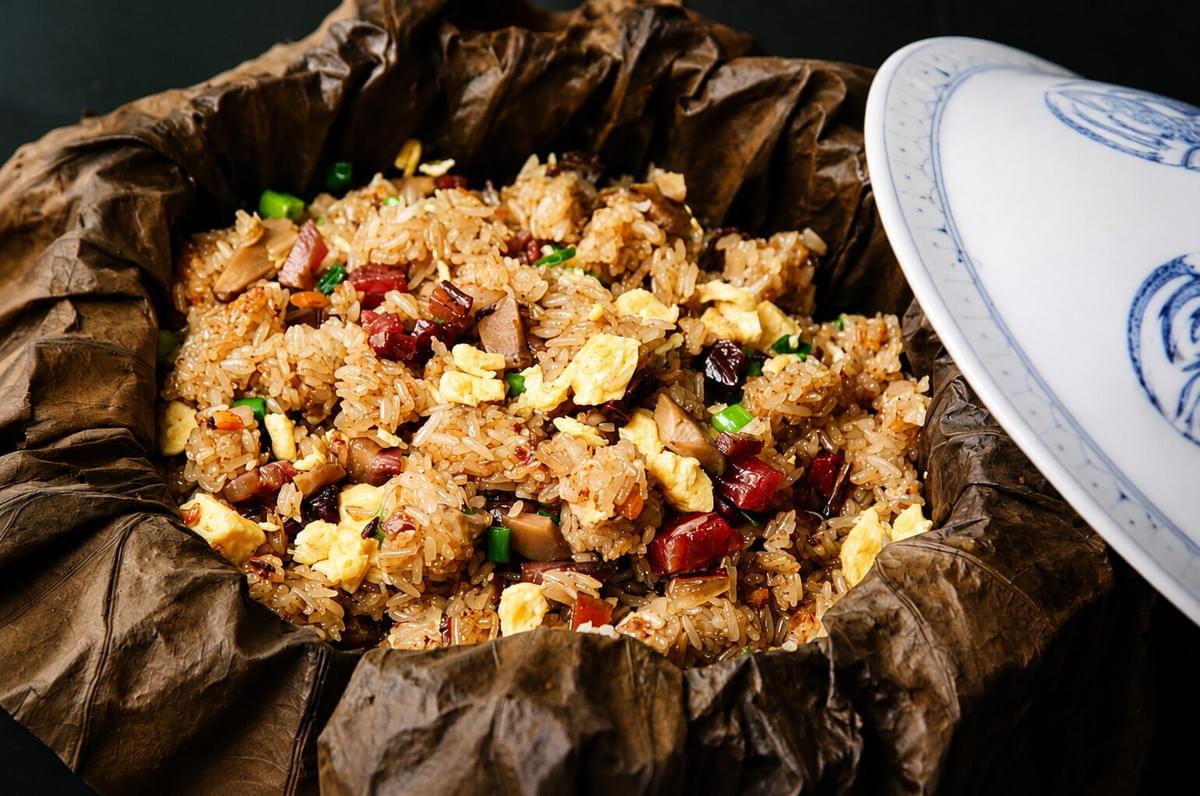 A close-up view of fried sticky rice with scrambled eggs, mushrooms, and Chinese sausage, served inside an open, crinkled lotus leaf with a white porcelain lid leaning against the side. A close-up view of fried sticky rice with scrambled eggs, mushrooms, and Chinese sausage, served inside an open, crinkled lotus leaf with a white porcelain lid leaning against the side.