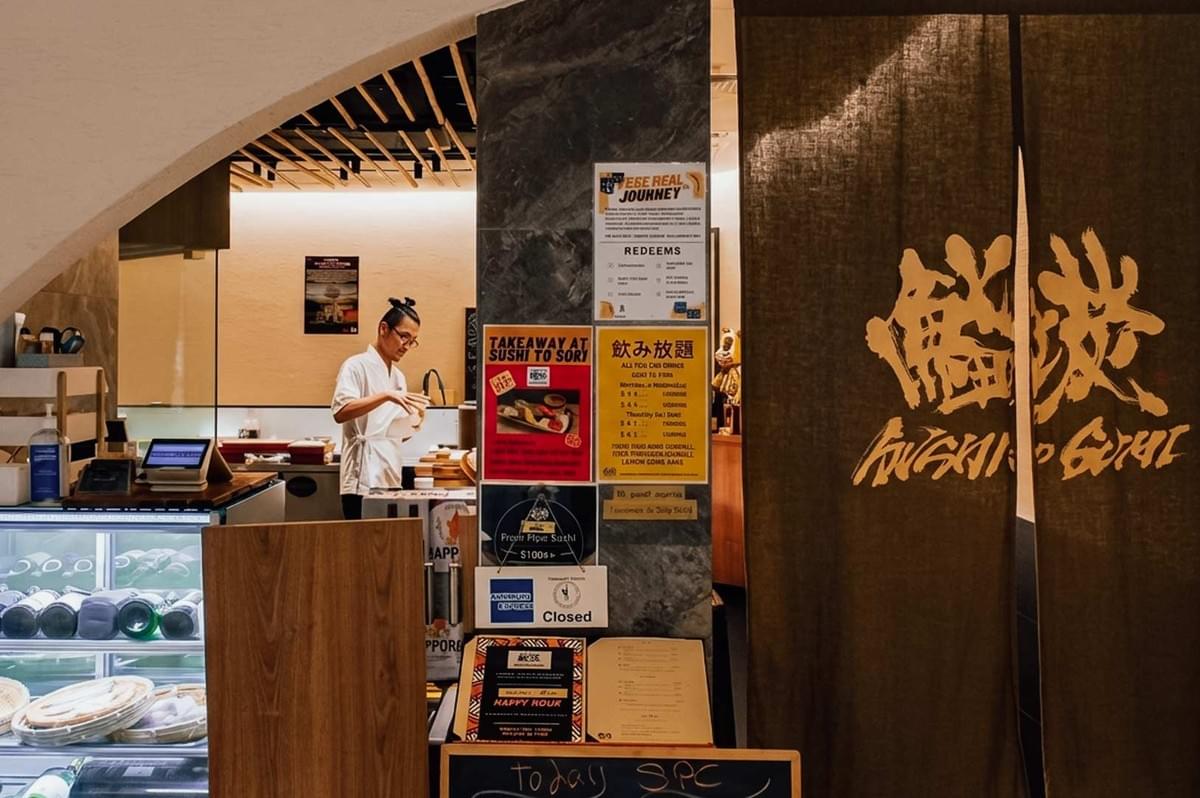 A chef working behind the counter seen through an archway at the entrance of "Sushi to Sumi," which is framed by a brown noren curtain displaying gold Japanese calligraphy on the right, alongside informational posters, a refrigerated display case, and a menu stand at the front. A chef working behind the counter seen through an archway at the entrance of "Sushi to Sumi," which is framed by a brown noren curtain displaying gold Japanese calligraphy on the right, alongside informational posters, a refrigerated display case, and a menu stand at the front.