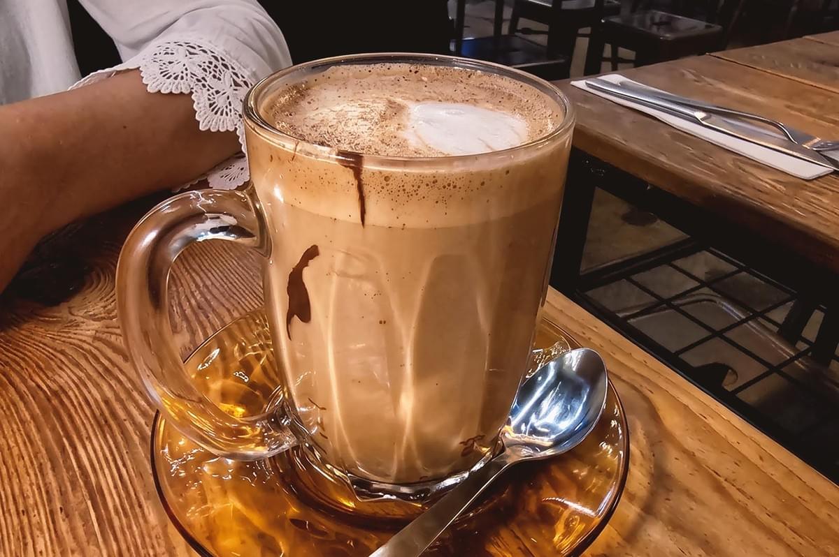 Close up of tall glass mug with creamy coffee with splashes of chocolate syrup placed in a glass plate along with a stirring spoon Close up of tall glass mug with creamy coffee with splashes of chocolate syrup placed in a glass plate along with a stirring spoon