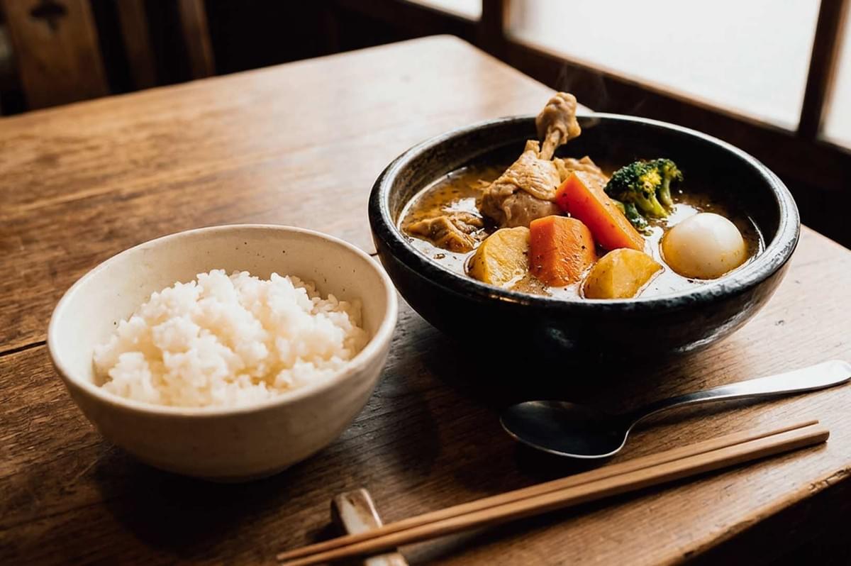 Foreground Items: A rustic dark ceramic bowl containing steaming Sapporo soup curry packed with a whole tender chicken leg, chunky pieces of potato, carrot, broccoli, and a soft-boiled egg. Next to it sits a white bowl filled with steamed white rice, a metal spoon, and wooden chopsticks resting on a chopstick holder. Foreground Items: A rustic dark ceramic bowl containing steaming Sapporo soup curry packed with a whole tender chicken leg, chunky pieces of potato, carrot, broccoli, and a soft-boiled egg. Next to it sits a white bowl filled with steamed white rice, a metal spoon, and wooden chopsticks resting on a chopstick holder.