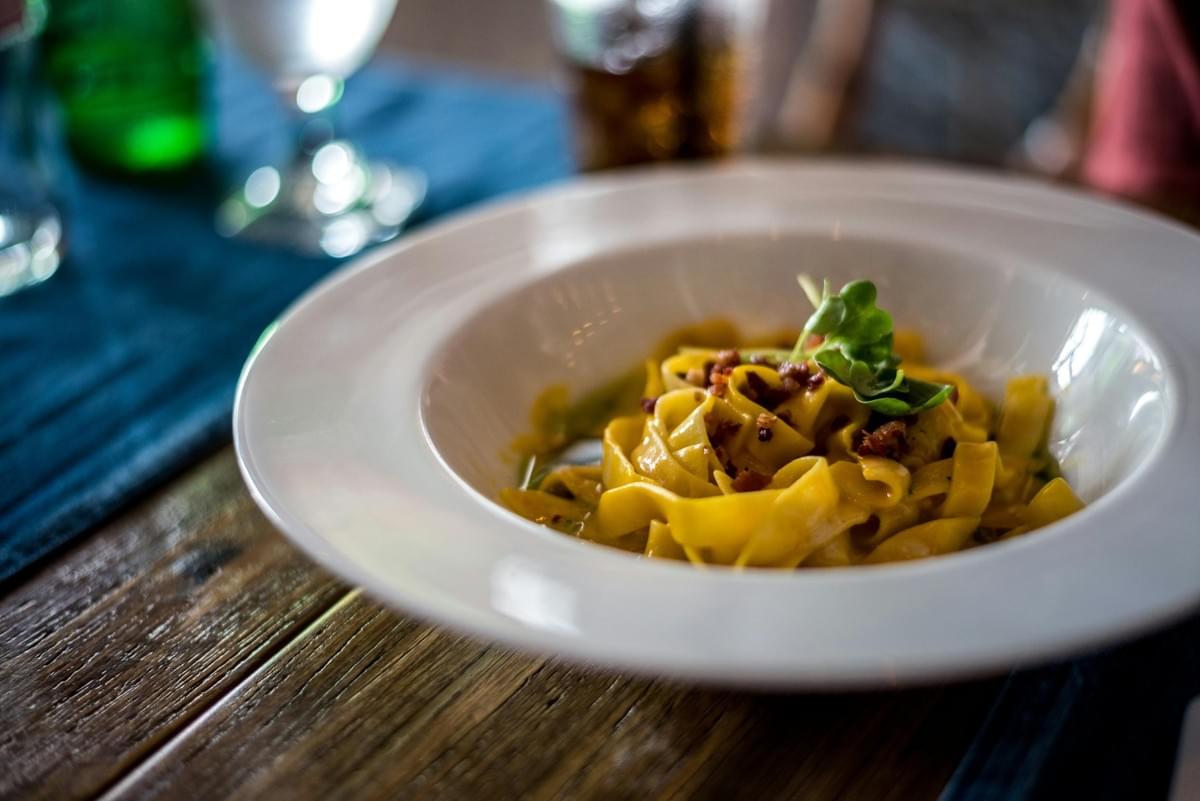 A wide white bowl filled with golden fettuccine pasta rests on a rustic wooden table. The dish is sprinkled with crispy pancetta bits and garnished with a fresh green leaf, set against a softly blurred restaurant background. A wide white bowl filled with golden fettuccine pasta rests on a rustic wooden table. The dish is sprinkled with crispy pancetta bits and garnished with a fresh green leaf, set against a softly blurred restaurant background.