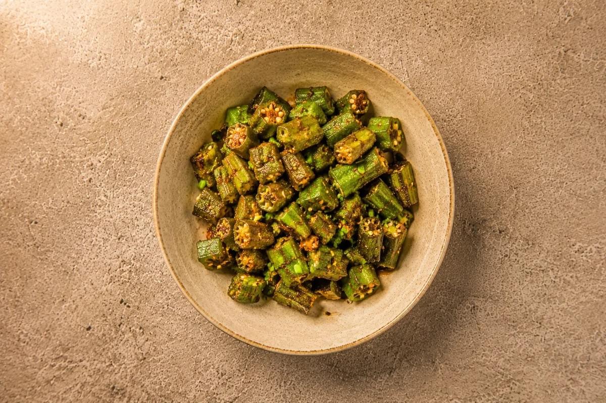 A top-down shot of seasoned, roasted okra pieces cut into bite-sized segments and served in a light beige ceramic bowl, placed on a textured stone surface. A top-down shot of seasoned, roasted okra pieces cut into bite-sized segments and served in a light beige ceramic bowl, placed on a textured stone surface.