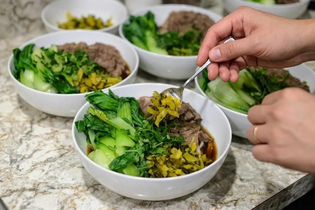 Multiple white bowls of Taiwanese beef noodle soup being garnished with pickled mustard greens and bok choy Multiple white bowls of Taiwanese beef noodle soup being garnished with pickled mustard greens and bok choy