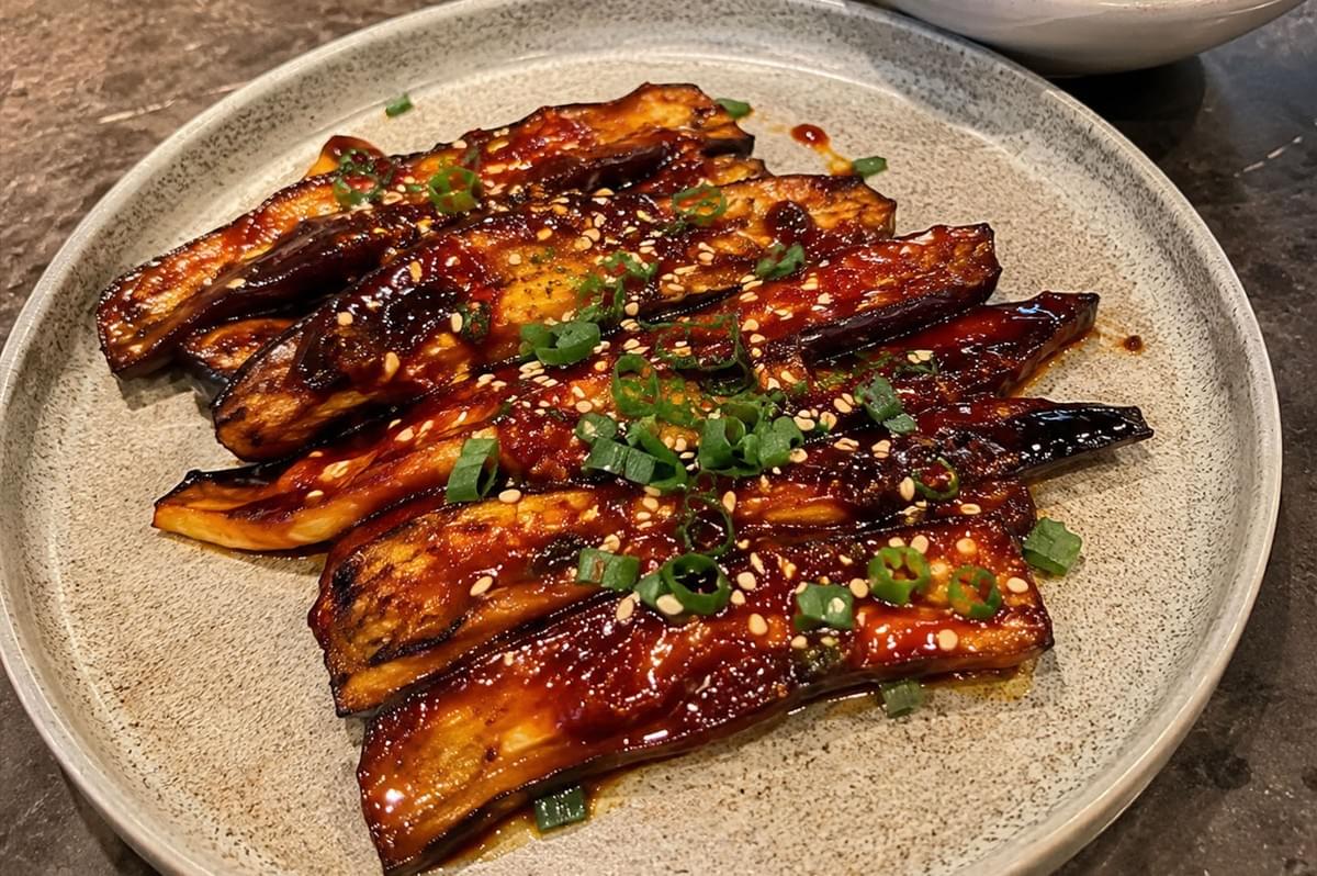A close-up, high-angle shot of a dish featuring long, tender slices of glazed eggplant arranged neatly on a speckled, light grey ceramic plate. The eggplant is coated in a thick, highly glossy, caramelized dark reddish-brown sauce, creating a sweet and savory appearance. The dish is garnished down the center with a sprinkle of toasted white sesame seeds and freshly chopped green scallions. In the upper-right background, a white ceramic bowl filled with steamed white rice is partially visible, all set against a dark, textured tabletop. A close-up, high-angle shot of a dish featuring long, tender slices of glazed eggplant arranged neatly on a speckled, light grey ceramic plate. The eggplant is coated in a thick, highly glossy, caramelized dark reddish-brown sauce, creating a sweet and savory appearance. The dish is garnished down the center with a sprinkle of toasted white sesame seeds and freshly chopped green scallions. In the upper-right background, a white ceramic bowl filled with steamed white rice is partially visible, all set against a dark, textured tabletop.