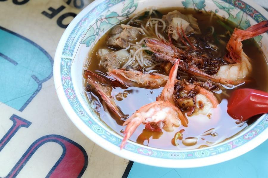 A top-down view shows a bowl of noodle soup filled with large cooked prawns, pork ribs, and fried shallots in a rich broth. The patterned ceramic bowl rests on a table next to a red plastic spoon. A top-down view shows a bowl of noodle soup filled with large cooked prawns, pork ribs, and fried shallots in a rich broth. The patterned ceramic bowl rests on a table next to a red plastic spoon.