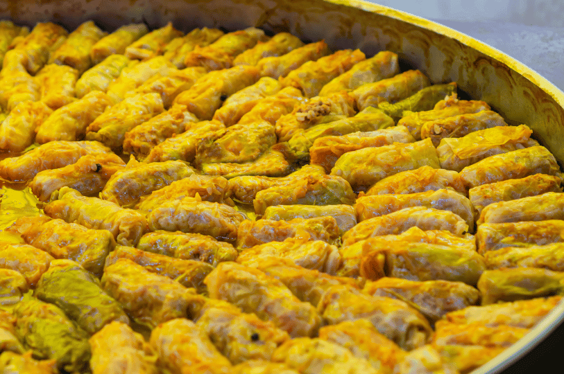 A large, deep pan filled edge-to-edge with a massive batch of freshly baked, golden-yellow Romanian cabbage rolls simmering in their juices. A large, deep pan filled edge-to-edge with a massive batch of freshly baked, golden-yellow Romanian cabbage rolls simmering in their juices.