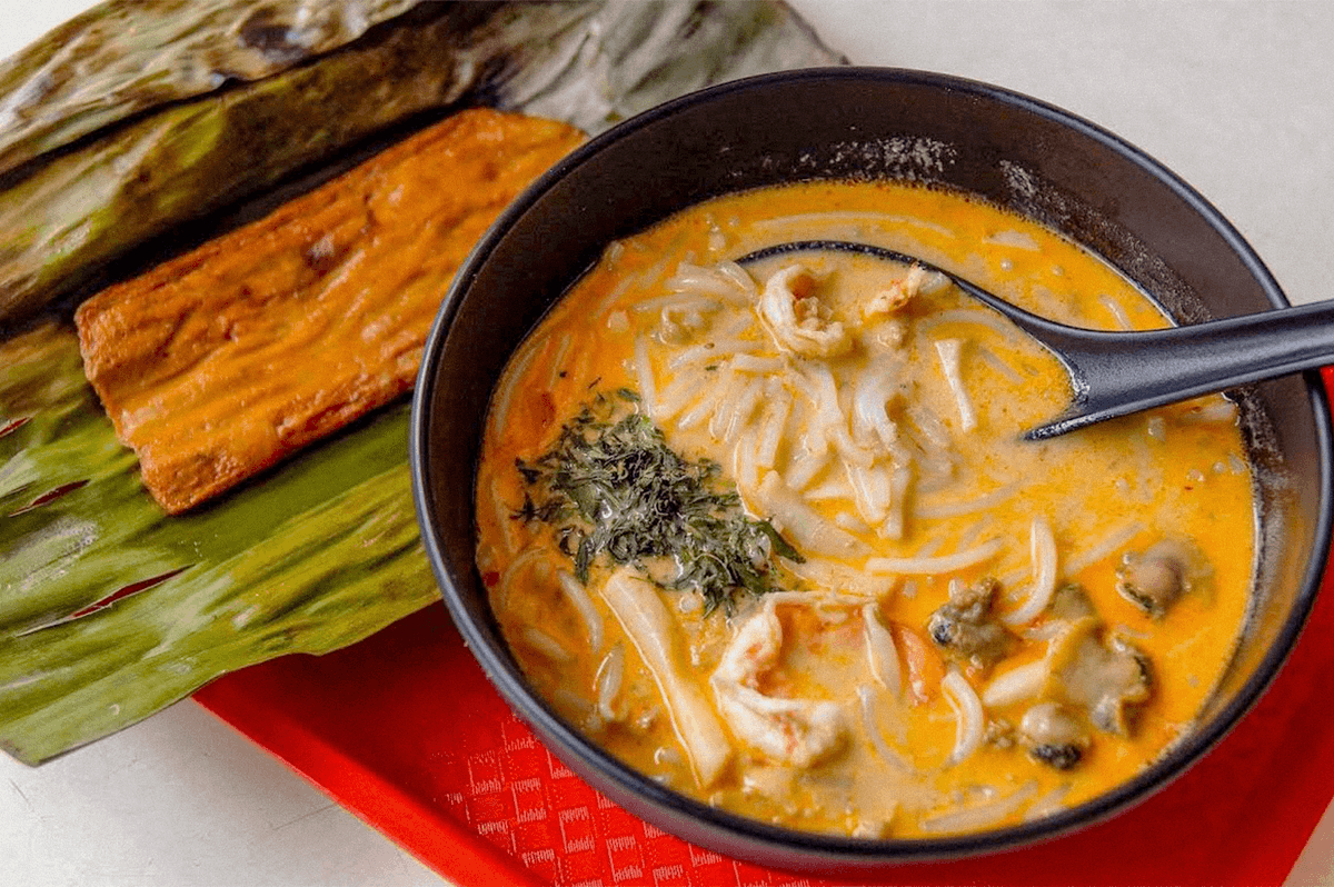 A bowl of creamy coconut noodle soup with seafood, served with grilled banana leaf-wrapped food. A bowl of creamy coconut noodle soup with seafood, served with grilled banana leaf-wrapped food.