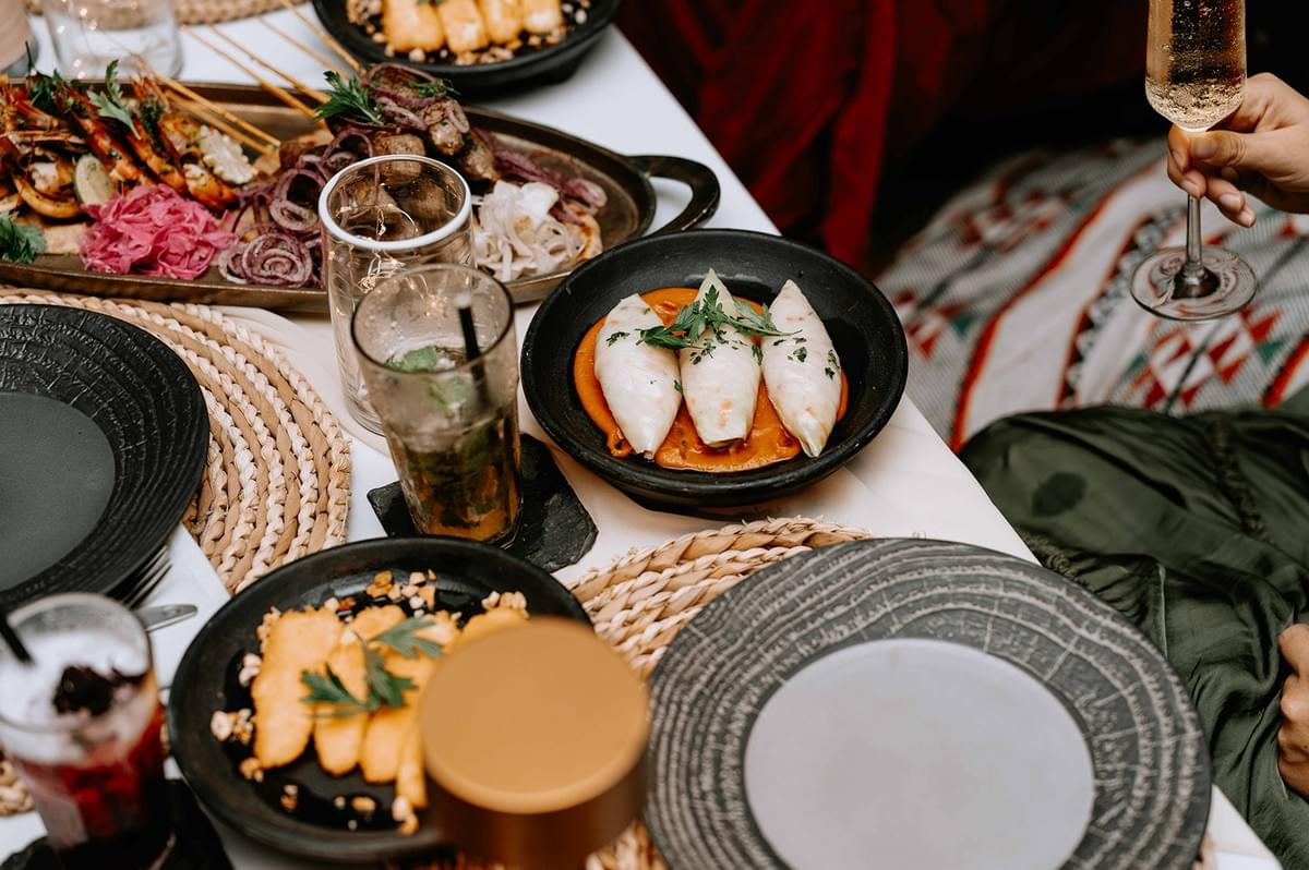 A top-down view of a shared table spread with gourmet food dishes, grilled skewers, stuffed squid, drinks, and woven placemats during a restaurant meal. A top-down view of a shared table spread with gourmet food dishes, grilled skewers, stuffed squid, drinks, and woven placemats during a restaurant meal.