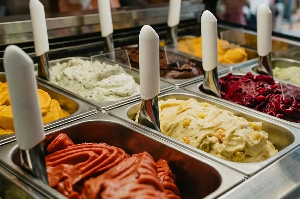 Assorted colorful gelato flavors in metal bins with white-handled scoops in a display case. Assorted colorful gelato flavors in metal bins with white-handled scoops in a display case.