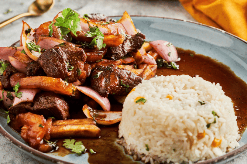 A modern plating of Lomo Saltado on a blue-grey plate. The beef and fries are tossed together in a glossy soy-based sauce, served with a perfectly molded dome of white rice and a sprinkle of cilantro. A modern plating of Lomo Saltado on a blue-grey plate. The beef and fries are tossed together in a glossy soy-based sauce, served with a perfectly molded dome of white rice and a sprinkle of cilantro.