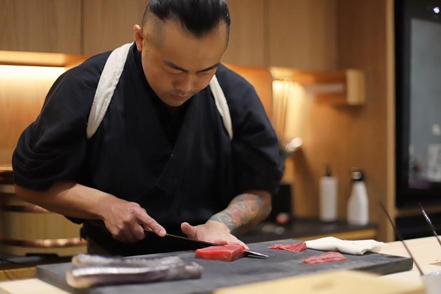 A sushi chef with a colorful sleeve tattoo carefully slices a piece of raw tuna on a dark prep board. He is dressed in a traditional black kimono, working in a warm, dimly lit kitchen with modern wooden accents. A sushi chef with a colorful sleeve tattoo carefully slices a piece of raw tuna on a dark prep board. He is dressed in a traditional black kimono, working in a warm, dimly lit kitchen with modern wooden accents.