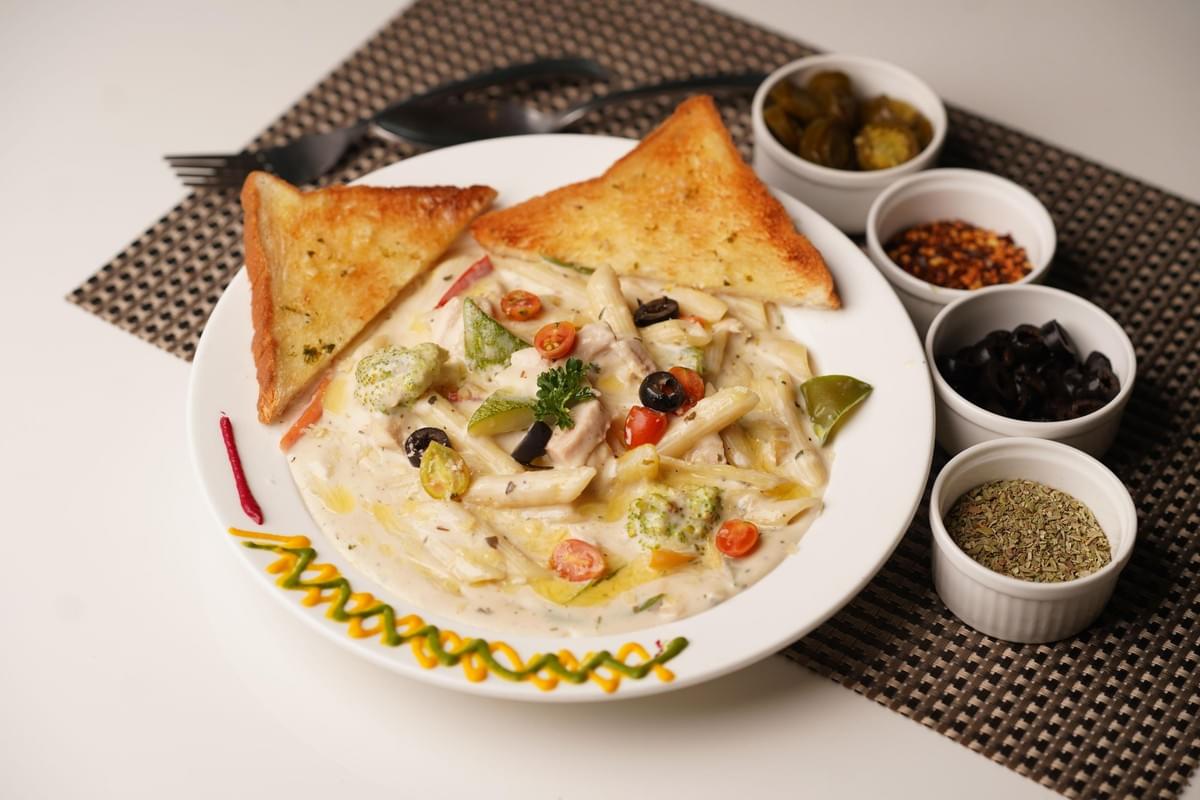 A large plate of creamy penne white sauce pasta is served with two triangular slices of golden garlic bread. Four small white condiment bowls containing sliced jalapenos, red chili flakes, black olives, and dried herbs sit alongside the plate on a checkered placemat. A large plate of creamy penne white sauce pasta is served with two triangular slices of golden garlic bread. Four small white condiment bowls containing sliced jalapenos, red chili flakes, black olives, and dried herbs sit alongside the plate on a checkered placemat.