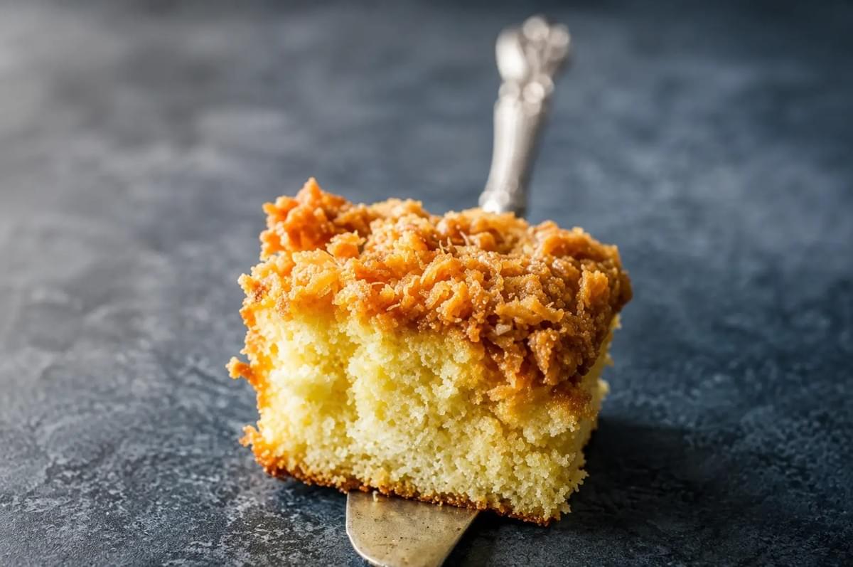 A single slice of yellow cake topped with caramelized coconut, held up on a metal dessert server. A single slice of yellow cake topped with caramelized coconut, held up on a metal dessert server.