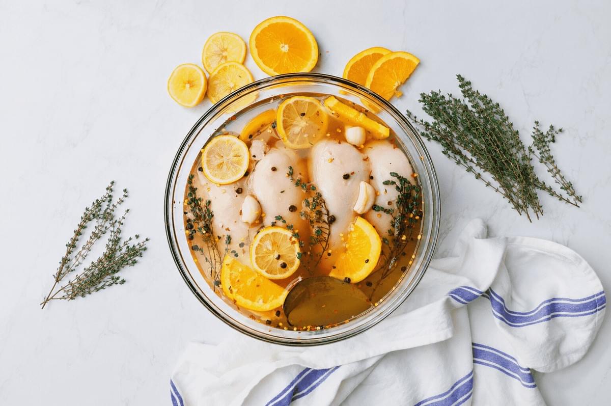 An overhead view of raw chicken breasts marinating in a glass bowl filled with liquid, lemon and orange slices, garlic cloves, peppercorns, and fresh herbs, arranged on a white marble countertop with loose citrus slices and thyme sprigs. An overhead view of raw chicken breasts marinating in a glass bowl filled with liquid, lemon and orange slices, garlic cloves, peppercorns, and fresh herbs, arranged on a white marble countertop with loose citrus slices and thyme sprigs.