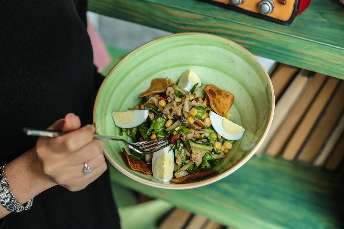 A person holds a large green bowl filled with a fresh salad topped with hard-boiled egg wedges, corn, and crispy chips. They use a fork to take a bite, set against a backdrop of a green wooden bookshelf. A person holds a large green bowl filled with a fresh salad topped with hard-boiled egg wedges, corn, and crispy chips. They use a fork to take a bite, set against a backdrop of a green wooden bookshelf.