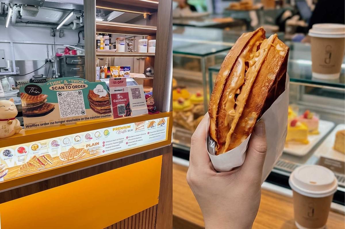 Left image alt text: A waffle kiosk with a wooden counter and a bright yellow front panel. A sign above the counter displays "Thin" and "Thick" waffle options with a QR code for ordering. The menu below shows various waffle flavors, while shelves behind the counter hold ingredients and supplies. A small lucky cat figurine sits on the left side of the counter. Right image alt text: A hand holding a freshly made waffle sandwich filled with a thick, creamy peanut butter-like filling in a white paper sleeve. The background features a softly blurred bakery display case with colorful cakes and pastries, along with takeaway coffee cups on the counter. Left image alt text: A waffle kiosk with a wooden counter and a bright yellow front panel. A sign above the counter displays "Thin" and "Thick" waffle options with a QR code for ordering. The menu below shows various waffle flavors, while shelves behind the counter hold ingredients and supplies. A small lucky cat figurine sits on the left side of the counter. Right image alt text: A hand holding a freshly made waffle sandwich filled with a thick, creamy peanut butter-like filling in a white paper sleeve. The background features a softly blurred bakery display case with colorful cakes and pastries, along with takeaway coffee cups on the counter.