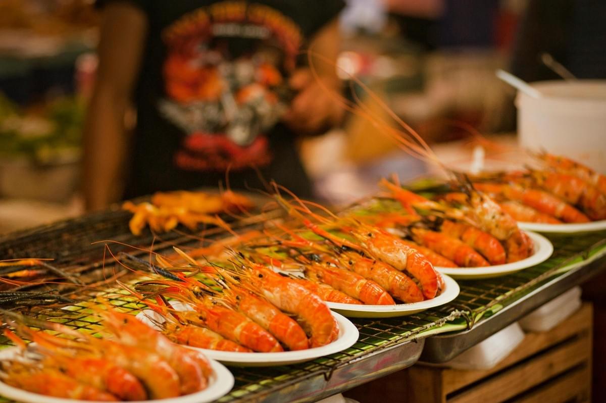 Large grilled prawns are neatly arranged on white plates atop a metal wire rack at an outdoor market. In the background, a person in a black t-shirt stands near the stall where the seafood is being prepared. Large grilled prawns are neatly arranged on white plates atop a metal wire rack at an outdoor market. In the background, a person in a black t-shirt stands near the stall where the seafood is being prepared.
